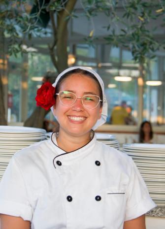 Chef sorridente con fiore rosso nei capelli, davanti a piatti impilati.