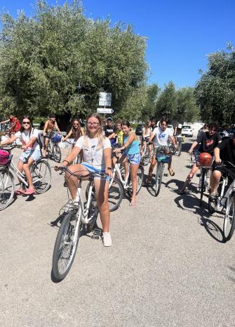 Gruppo di giovani in bici sotto il sole, tra alberi verdi.