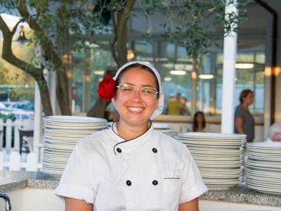 Chef sorridente con fiore rosso nei capelli, davanti a piatti impilati.