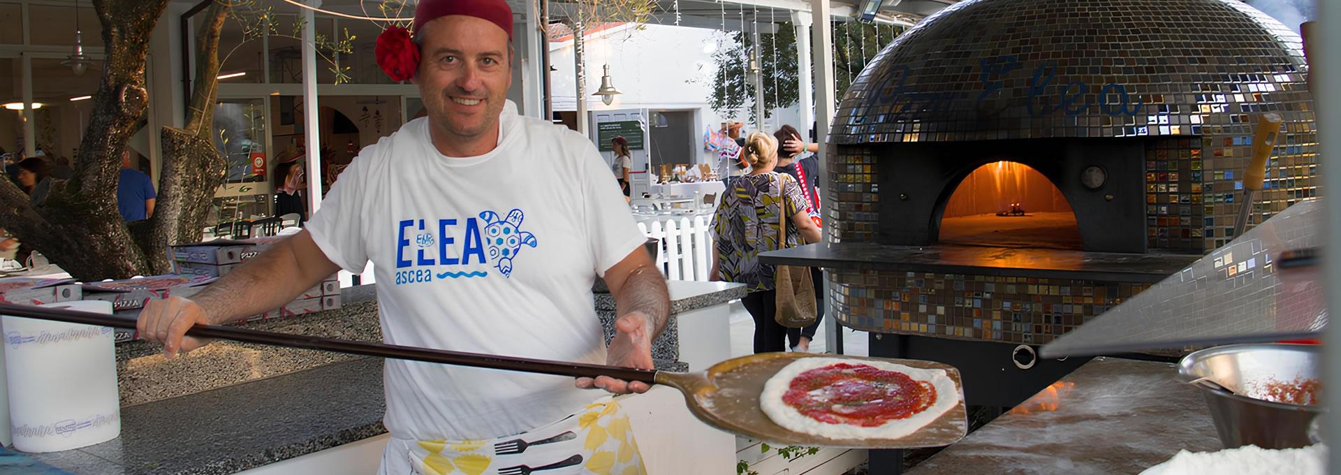 Pizzaiolo sorridente con pizza cruda, forno a legna scintillante sullo sfondo.