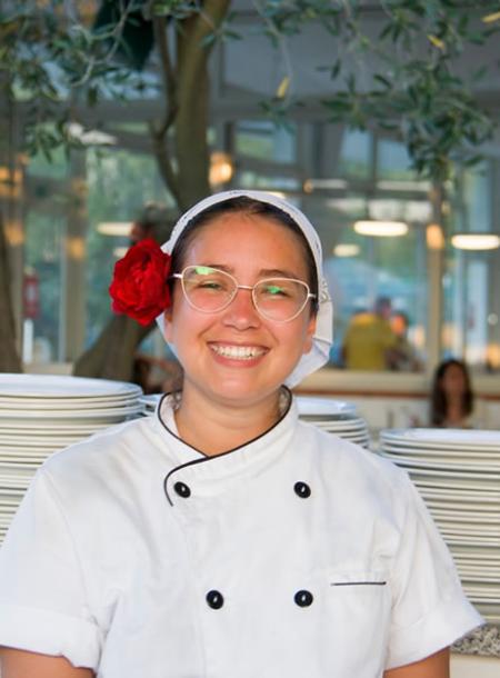 Chef sorridente con fiore rosso tra i capelli, atmosfera accogliente e luminosa.