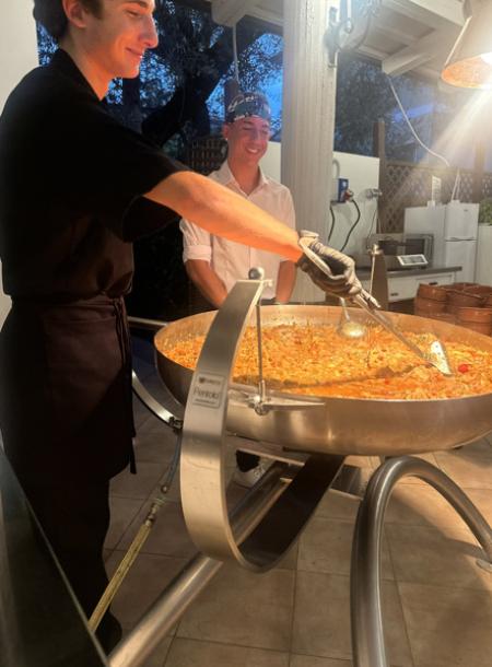 Cuochi sorridenti mescolano un'enorme pentola di cibo, atmosfera calda e conviviale.